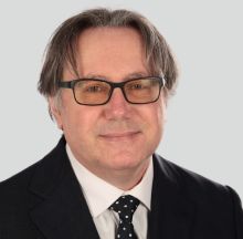 Ritratto professionale di Massimo Dalla Mora, architetto, con giacca, camicia bianca e cravatta a pois, sfondo grigio neutro.