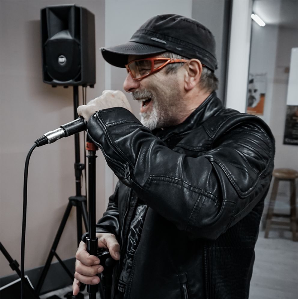 Cantante con cappellino e giacca di pelle che canta al microfono su asta in sala prove; scatto reportage per il portfolio di Natalia Canaletti